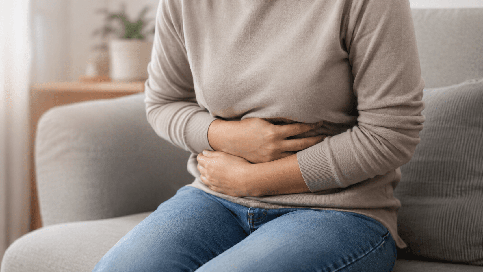 Person sitting on a sofa holding their abdomen with both hands, appearing to experience stomach pain.